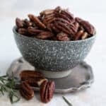 Salted pecans with rosemary piled up in a speckled gray bowl on a pewter plate.