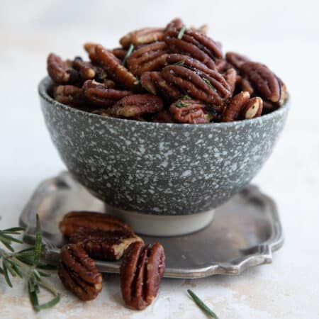 Salted pecans with rosemary piled up in a speckled gray bowl on a pewter plate.
