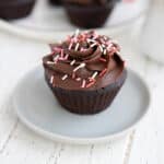 A keto chocolate cupcake with chocolate frosting and sprinkles on a small gray plate on a white wooden table.
