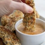 A hand dipping a chicken tender made with everything bagel seasoning into a small white bowl with honey mustard.