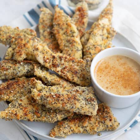 Keto chicken tenders with everything bagel seasoning piled up on a white plate over a blue and white striped napkin.