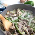 A wooden spoon digging into a pan full of Philly Cheesesteak fillings covered in melted cheese.