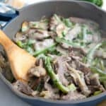 A wooden spoon digging into a pan full of Philly Cheesesteak fillings covered in melted cheese.