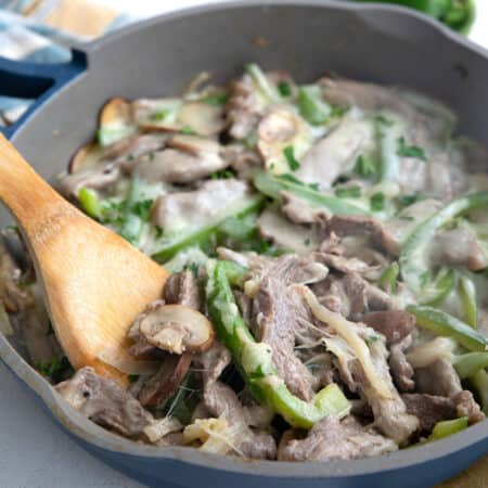 A wooden spoon digging into a pan full of Philly Cheesesteak fillings covered in melted cheese.