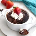 Keto chocolate protein mug cake in a white ramekin with whipped cream and strawberries on top. Several spoonfuls of cake have been taken out and one sits in front of the ramekin on a small metal spoon.