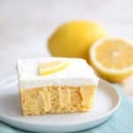 A slice of Keto Lemon Poke Cake on a white plate with a whole lemon and half a lemon in the background.