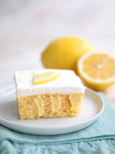 A slice of Keto Lemon Poke Cake on a white plate with a whole lemon and half a lemon in the background.
