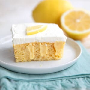 A slice of Keto Lemon Poke Cake on a white plate with a whole lemon and half a lemon in the background.