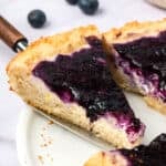 A slice of Keto Blueberry Cream Cheese Coffee Cake being pulled away from the rest of the cake on a white platter.