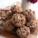 A pile of Keto Brown Butter Pecan Muffins on a wooden cutting board with a vase of flowers behind it.