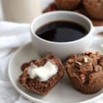 Two Keto Brown Butter Pecan Muffins sit on a plate with a cup of coffee. One muffin is broken in half, and has butter melting on it.