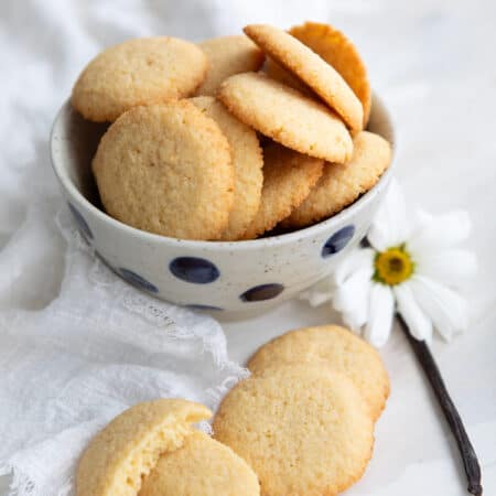 Vertical image of Keto Vanilla Wafers piled up in a polka dot bowl with a vanilla bean and a white flower beside it.
