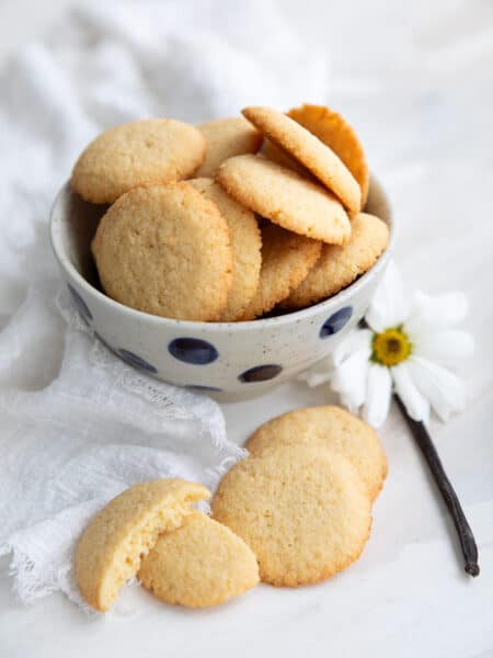 Vertical image of Keto Vanilla Wafers piled up in a polka dot bowl with a vanilla bean and a white flower beside it.