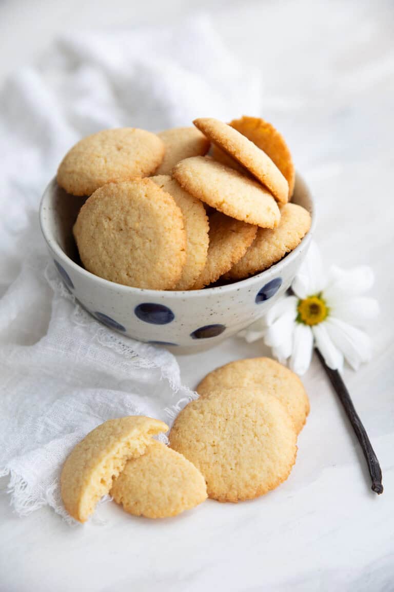 Vertical image of Keto Vanilla Wafers piled up in a polka dot bowl with a vanilla bean and a white flower beside it.