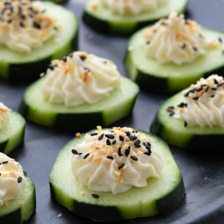 Cucumber slices topped with cream cheese and everything bagel seasoning on a black plate on a white table top.