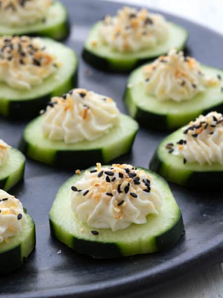 Cucumber slices topped with cream cheese and everything bagel seasoning on a black plate on a white table top.