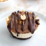 Close up shot of a mini keto cheesecake with chocolate crust, caramel, peanuts, and chocolate drizzle on a white plate.