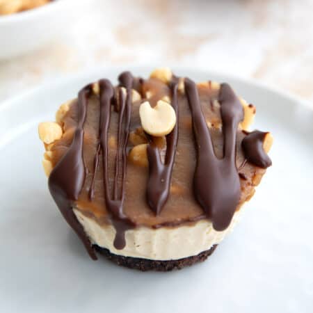 Close up shot of a mini keto cheesecake with chocolate crust, caramel, peanuts, and chocolate drizzle on a white plate.