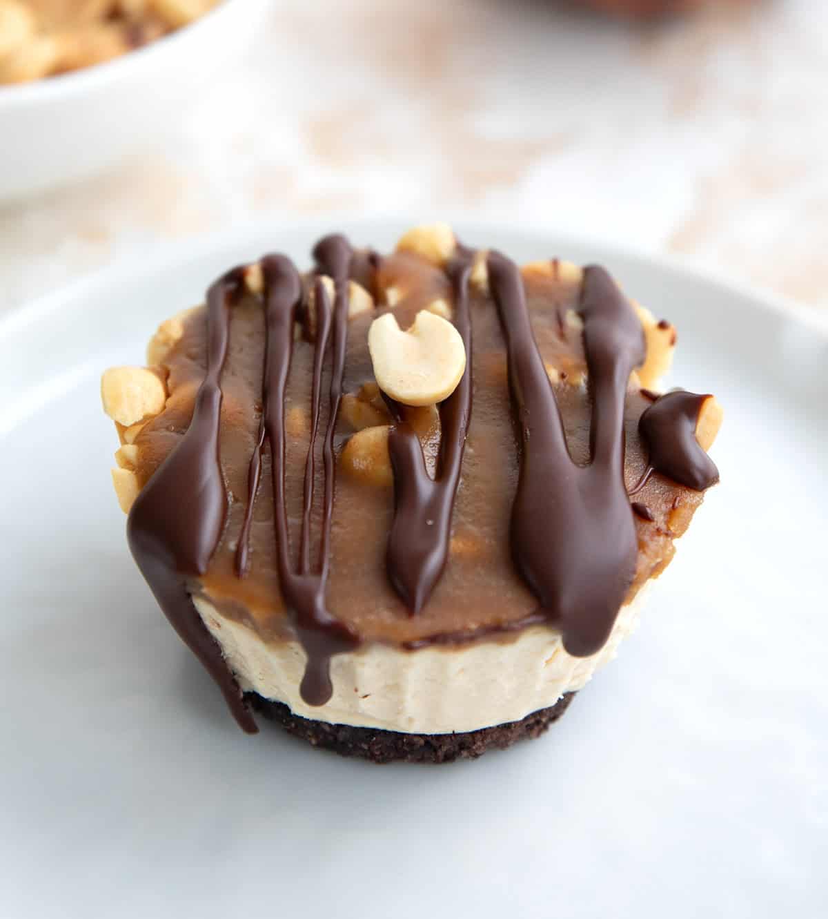 Close up shot of a mini keto cheesecake with chocolate crust, caramel, peanuts, and chocolate drizzle on a white plate.