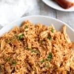 Titled image of Keto BBQ Shredded Chicken piled up in a white bowl on a gray wooden table.