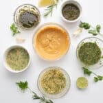 Top down image of 6 keto friendly chicken marinades on a white background, surrounded by sprigs of parsley, rosemary and cilantro, cloves of garlic, a lemon slice and a lime slice.