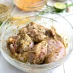 Skinless chicken thighs in a glass bowl coated in a chimichurri marinade on a white wooden table.