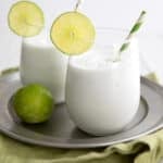 Two glasses of keto coconut limeade on a round metal plate over a green napkin, all on a white wooden table top.