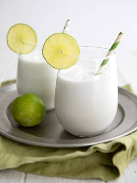 Two glasses of keto coconut limeade on a round metal plate over a green napkin, all on a white wooden table top.