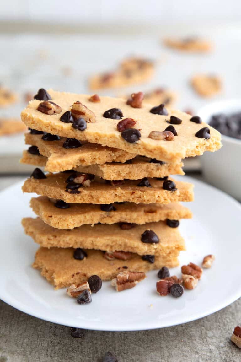 Pieces of keto cookie brittle piled up on a white plate on a gray concrete table.