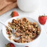 A spoon digging into a small white bowl filled with Keto Air Fryer Granola, with strawberries arranged around it.