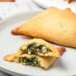 Keto Spanakopita arranged on a white plate with one of the pastries broken open to show the inside.