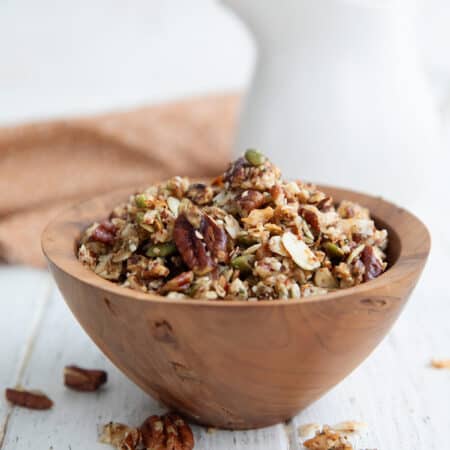 Keto Air Fryer Granola piled up in a wooden bowl on a white wooden table, with a jug of milk in the background.