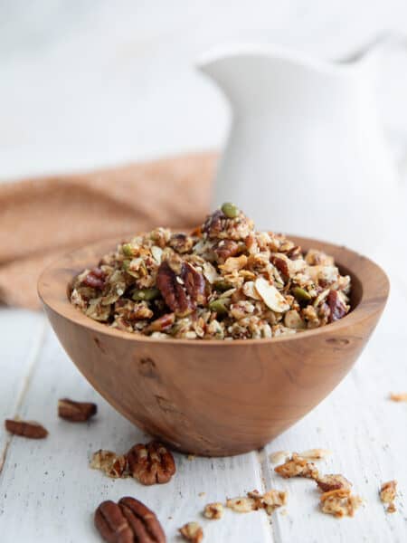 Keto Air Fryer Granola piled up in a wooden bowl on a white wooden table, with a jug of milk in the background.