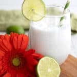 A glass of keto coconut limeade on a small wooden cutting boars with half a lime and a coral colored gerbera daisy.