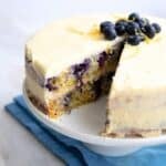 Keto Lemon Blueberry Cake with a slice cut out of it, on a white cake stand over a blue napkin.