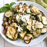 Grilled zucchini salad with feta and basil piled up in a white bowl on a white wooden table.