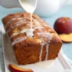 Vanilla glaze being drizzled over a loaf of keto peach bread on a white platter.