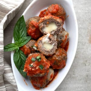 Top down image of keto stuffed meatballs with tomato sauce piled up in a white oval dish. One meatball is cut in half to show the inside.