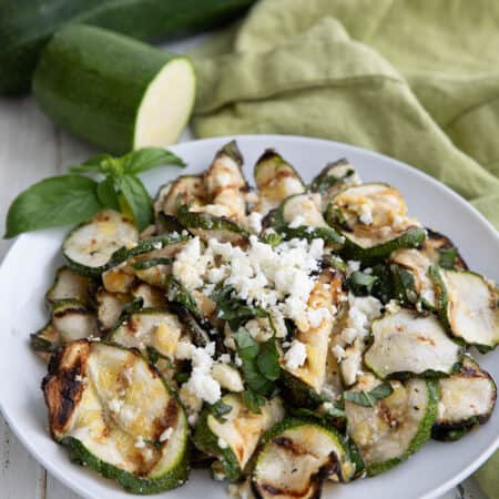 A white bowl filled with grilled zucchini and feta, on a white wooden table, with zucchini in the background.