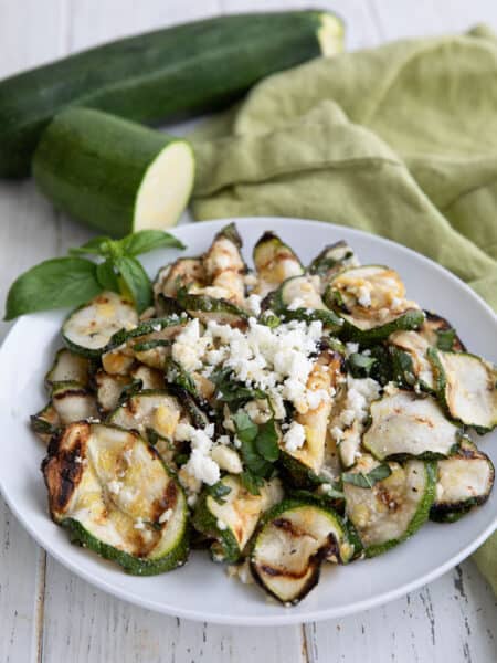 A white bowl filled with grilled zucchini and feta, on a white wooden table, with zucchini in the background.