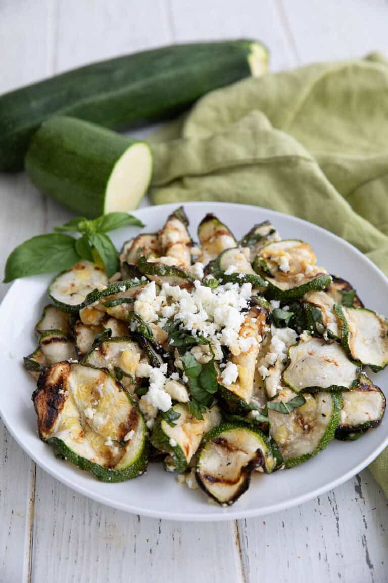 A white bowl filled with grilled zucchini and feta, on a white wooden table, with zucchini in the background.