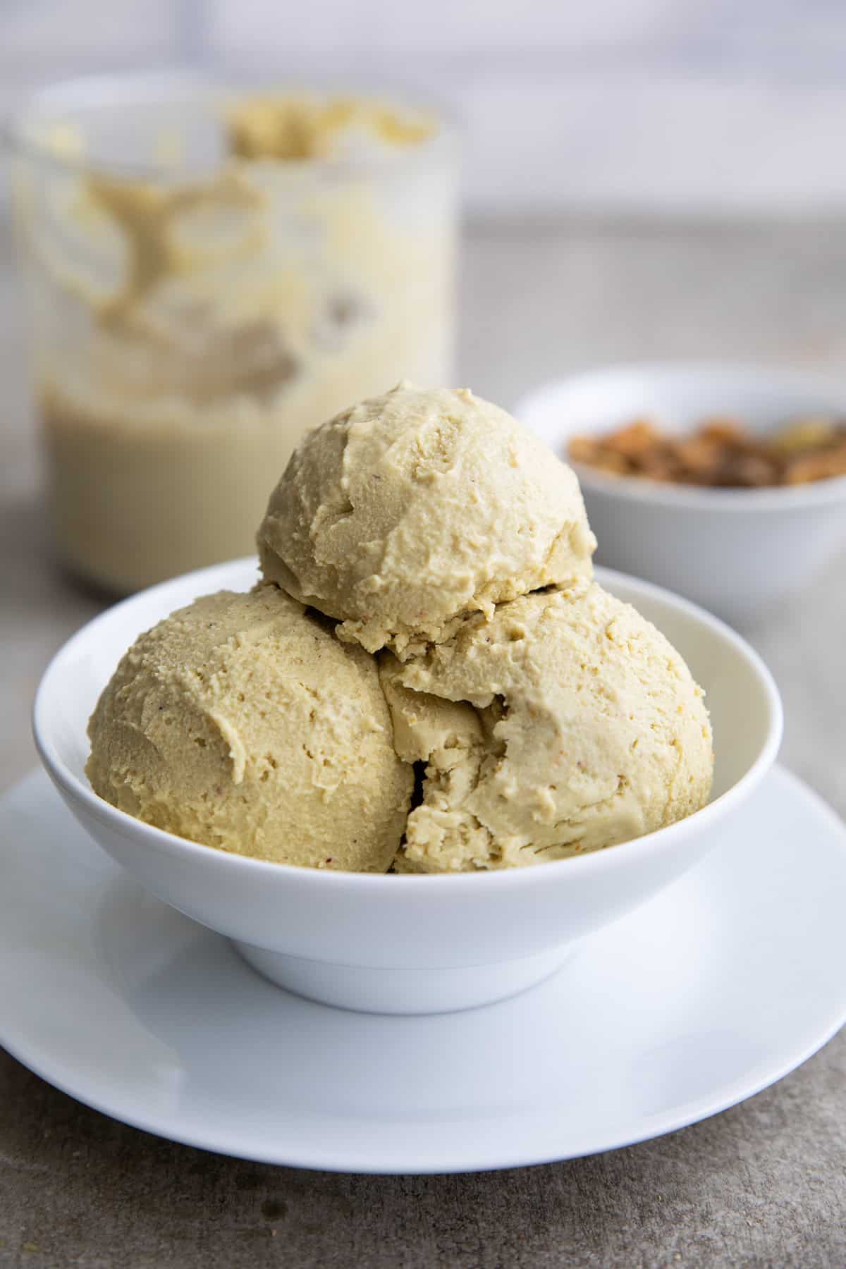 A white bowl with three scoops of keto pistachio ice cream, with a bowl of pistachios and the ice cream container in the background.