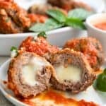 Three air fryer stuffed meatballs covered with tomato sauce on a white plate. One of the meatballs is cut in half to show the cheese inside.