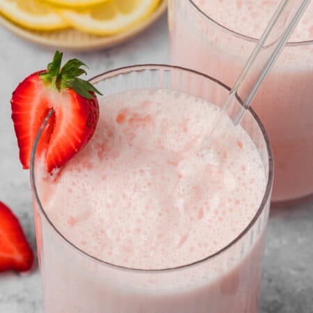 Close up shot of a glass of keto strawberry frosted lemonade with half a strawberry on the edge and a glass straw.
