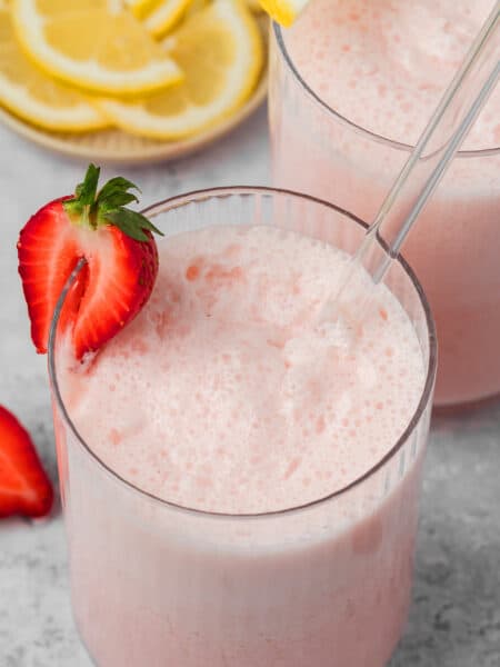 Close up shot of a glass of keto strawberry frosted lemonade with half a strawberry on the edge and a glass straw.