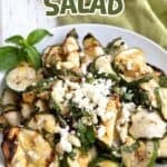 Titled image of a white bowl filled with grilled zucchini and feta, on a white wooden table.