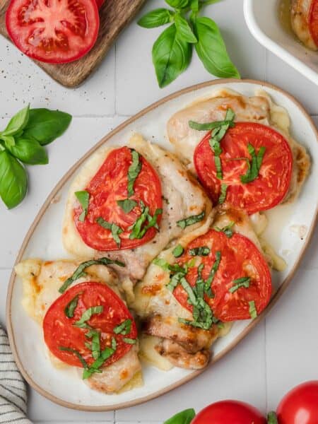 Top down image of cooked chicken thighs topped with melted mozzarella, sliced tomatoes, and chopped basil on a white oval platter with a brown rim.