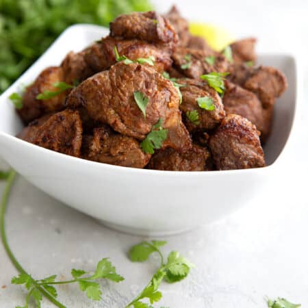 A white bowl filled with Air Fryer Steak Bites on a gray table, with a sprig of cilantro in the foreground.