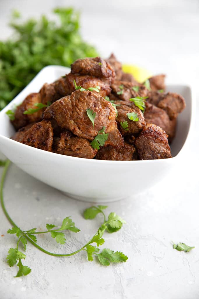 A white bowl filled with Air Fryer Steak Bites on a gray table, with a sprig of cilantro in the foreground.