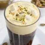 Titled image of a tall glass of iced coffee topped with keto pistachio cream cold foam, with chopped pistachios on top.